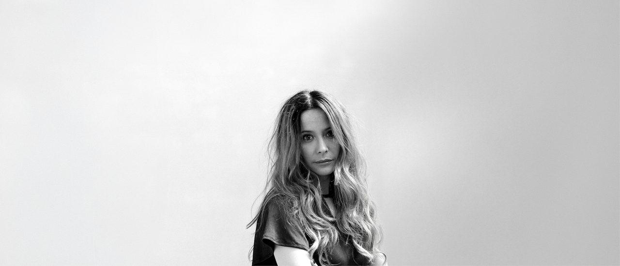 A black and white photo of a woman with long blonde hair cascading down past her shoulders, wearing a short-sleeved black dress. She looks at the camera with a slight smile.