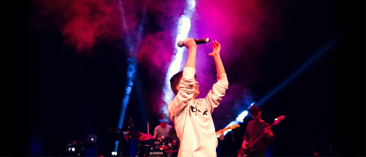 A young boy wearing a white hoodie raises his arms aloft, with a microphone in his left hand. A drummer and guitarist are playing behind him, illuminated by smoky pink lights and white lasers.