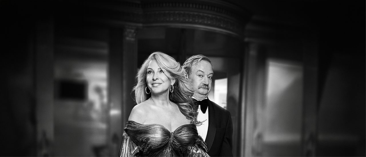 A black and white photo of a glamorous blonde woman in an off-the-shoulder pleated lurex dress and big gold hoop earrings, with a confident smile. Behind her a man with a moustache, wearing formal black tie, looks towards the camera with an exasperated expression.