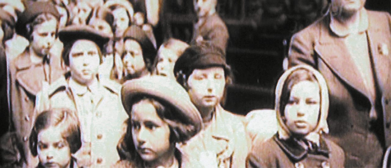 A grainy historical black and white photo of a group of children in the 1930s, dressed in coats and hats, some wearing labels around their necks and looking anxious and uncertain.
