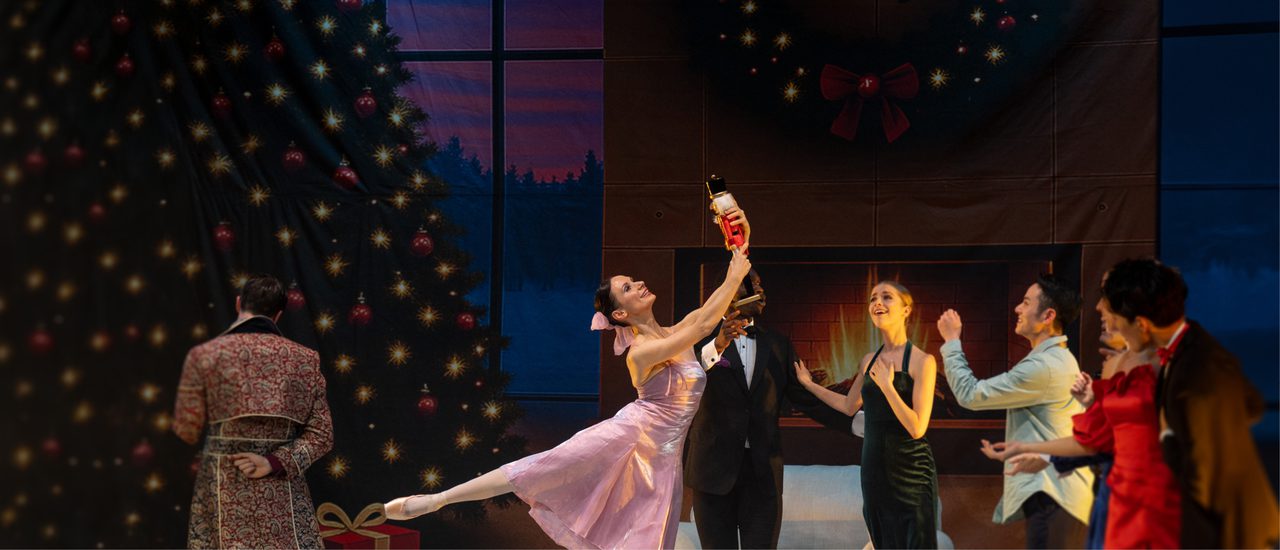 A female dancer wearing a pink satin party dress, her dark hair fastened in a bun with a pink bow, performs an arabesque. She's holding a Nutcracker doll aloft. Other party guests admire the toy. Behind them is a decorated Christmas tree, a roaring fire, white armchair and some wrapped gift boxes.