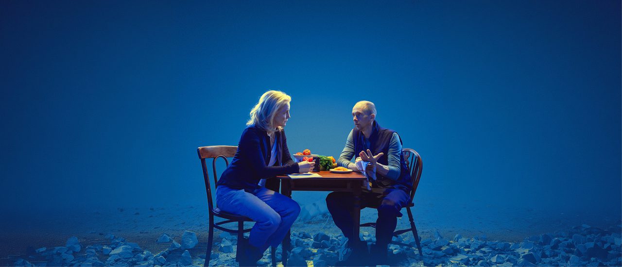 A blonde woman and a short-haired man sit looking at each other, in serious conversation, on either side of a table on which are bowls and plates of tomatoes and other fruit and vegetables. She has a notebook. They are illumined against a blue background and the ground is covered in shattered rocks.