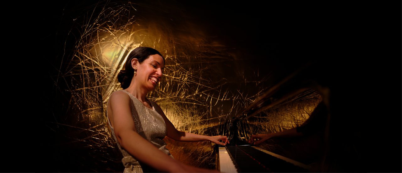 A smiling woman, her dark hair tied back in a chignon, sits playing the piano. She wears a sleeveless, dull gold shimmering dress, and has an illuminated gold background behind her.