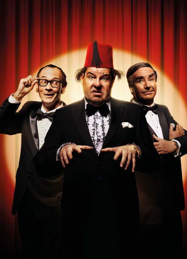 Three men stand together in front of a red curtain, lit with a golden spotlight. They all wear black tie. The man on the left wears glasses with thick black frames, which he holds up to his face with two fingers. He looks off to one side with a cheeky smile. The man in the centre wears a red fez and holds both hands out in front of him, eyes wide. The man on the right crosses his arms over his chest and raises one eyebrow with a slight smile.