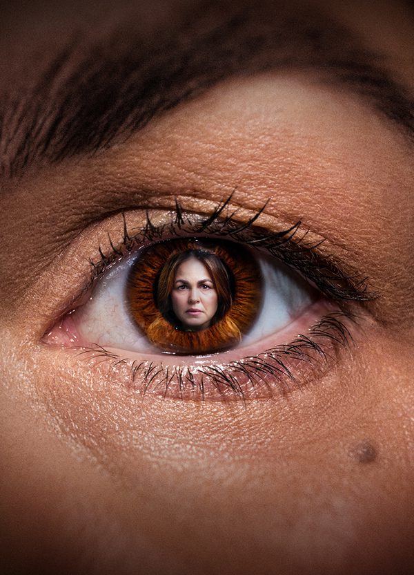 A close up of a woman's eye, within the pupil is an image of the same woman. She has dark brown hair and has a serious expression on her face.