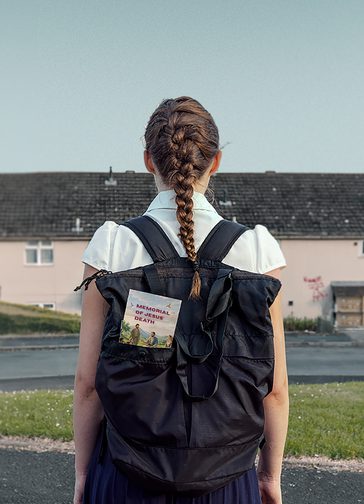 A young girl photographed from behind. She wears school uniform, a navy, pleated skirt and a short-sleeved, white blouse, and a black backpack. Her hair is tied back in a single, neat, French plait. A pamphlet which reads 'Memorial of Jesus' Death' can be seen poking out of her bag. A residential street with patches of grass, tarmac and terraced houses can be seen in the background, with a murky, grey sky.