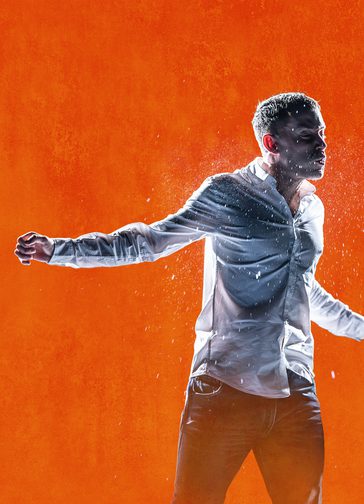 Against a bright orange background, a young man is in motion, as though mid-fight. He wears a white shirt. He holds one arm out to the side and his face looks pained. Water sprays around him.