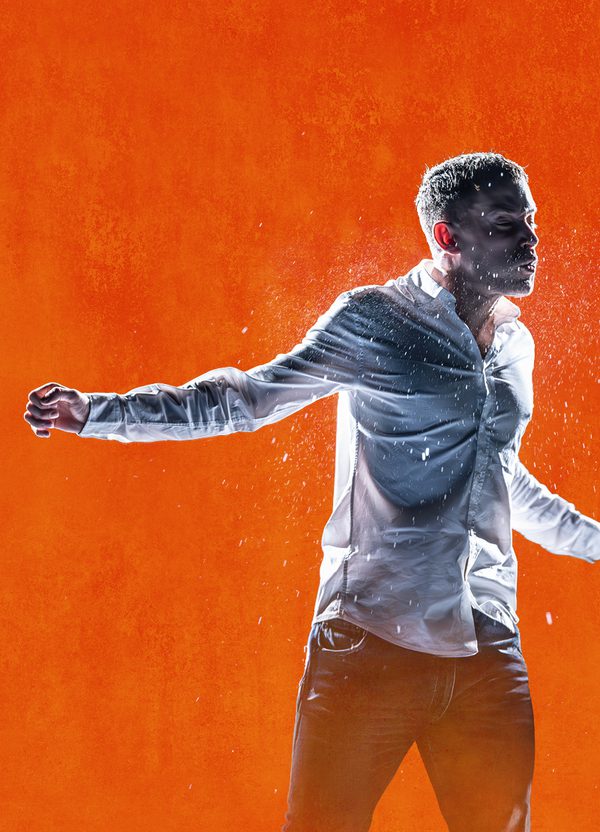 Against a bright orange background, a young man is in motion, as though mid-fight. He wears a white shirt. He holds one arm out to the side and his face looks pained. Water sprays around him.