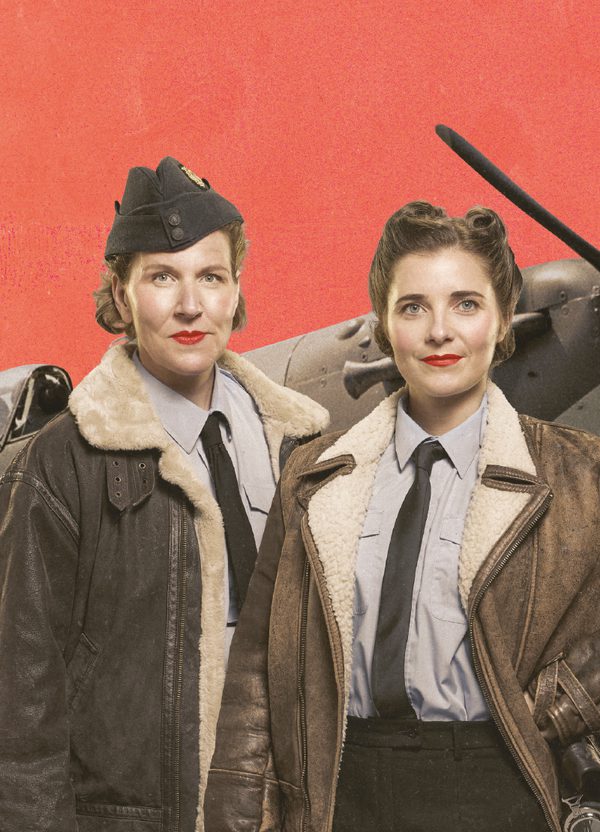 Two women in WWII RAF uniform, grey shirts, dark ties, brown aviator jackets and one in a dark hat, stand proudly together. They wear bright red lipstick and their hair is neatly styled in 1940s styles. They smile and look directly into the camera. Behind them is a spitfire plane against a coral background.