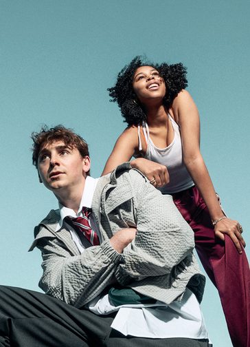 Two young people pose together against a light blue background. The boy crouches down, arms crossed over his chest, looking into the distance pensively. He wears school uniform and a thin bomber jacket. The girl stands, leaning on his shoulder with one arm and bending down with the other hand on her knee, looking up in the opposite direction with a smile. She wears burgundy, shiny tracksuit bottoms, a white vest top and hoop earrings.