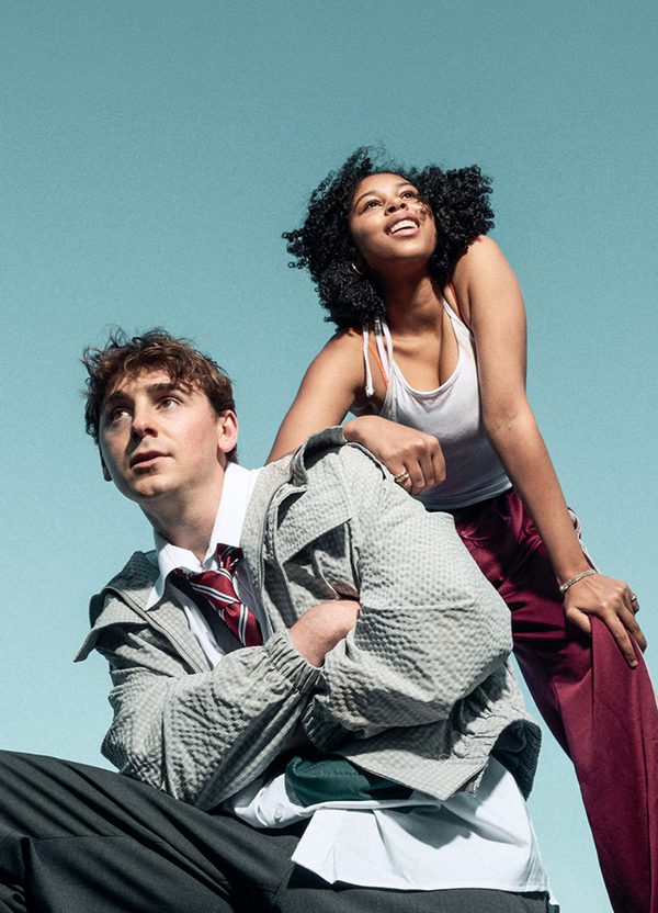 Two young people pose together against a light blue background. The boy crouches down, arms crossed over his chest, looking into the distance pensively. He wears school uniform and a thin bomber jacket. The girl stands, leaning on his shoulder with one arm and bending down with the other hand on her knee, looking up in the opposite direction with a smile. She wears burgundy, shiny tracksuit bottoms, a white vest top and hoop earrings.