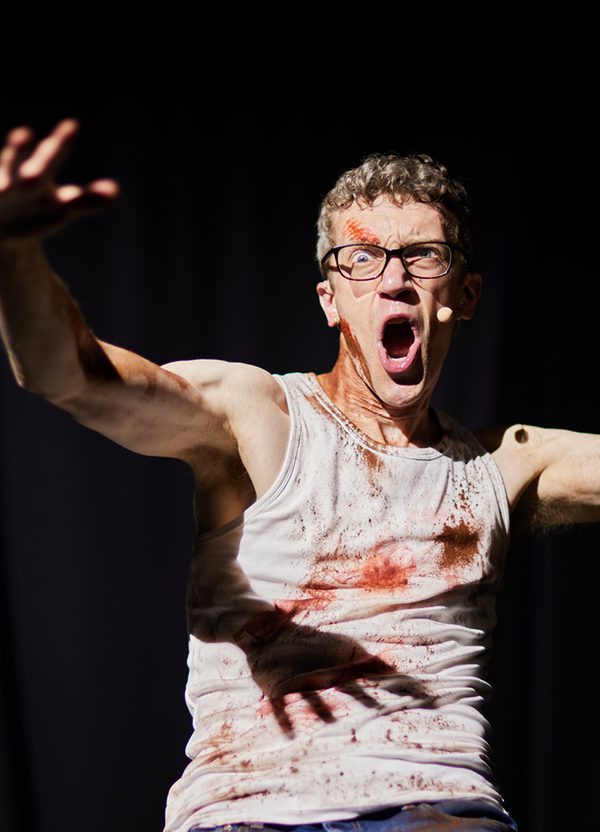 Against a black curtain, a man stands in a dramatic pose: his hands are raised with his fingers splayed out and he appears to be shouting, with one eye wide. He wears glasses and a white vest and is smeared with blood.