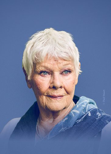 A photo of Dame Judi Dench on an indigo background. She looks off into the distance with a gentle smile. She has a blue scarf with white leaves draped over her shoulders.