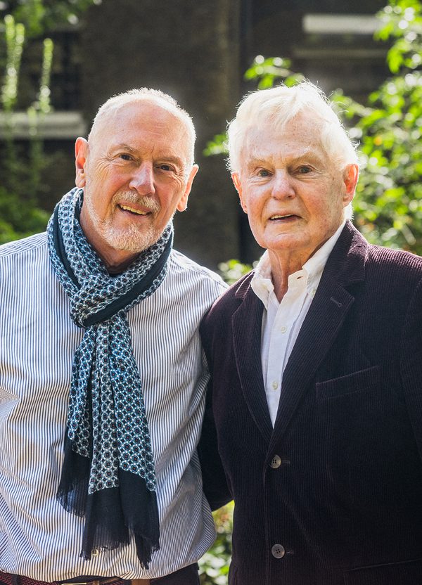 Two men (Derek Jacobi, right, and Richard Clifford, left) are stood side by side, an arm around each other. The man on the right (Derek Jacobi) is wearing a purple courdroy blazer and a white shirt, the man on the left (Richard Clifford) is wearing a blue striped shirt and a blue patterned scarf. They are both smiling at the camera.