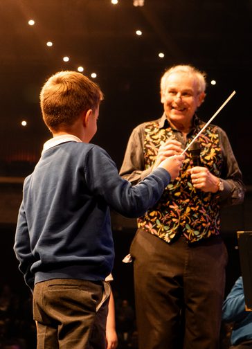 A small boy with his back to the camera stands holding a conductor's baton. An older man, wearing an embroidered waistcoat, stands smiling down at him.