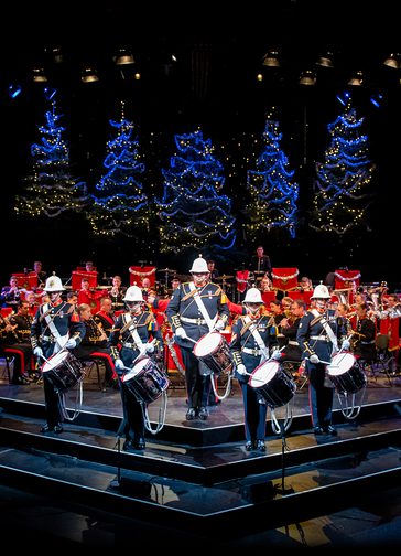 The Band of HM Royal Marines stand on the Festival Theatre stage dressed in military full dress, facing out front. Five drummers stand at the front, with the rest of the band seated behind. Behind them are five blue-lit