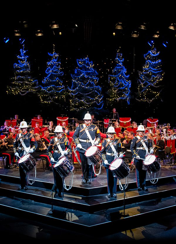 The Band of HM Royal Marines stand on the Festival Theatre stage dressed in military full dress, facing out front. Five drummers stand at the front, with the rest of the band seated behind. Behind them are five blue-lit