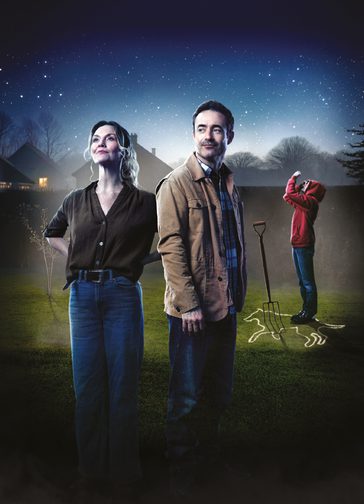 A casually dressed woman and man stand in a garden at night-time, against silhouetted houses, trees and a starry sky. Behind them, a young person in a red hoodie stands on an crime scene-type outline of a dog on the grass, with a pitchfork stuck into the ground.