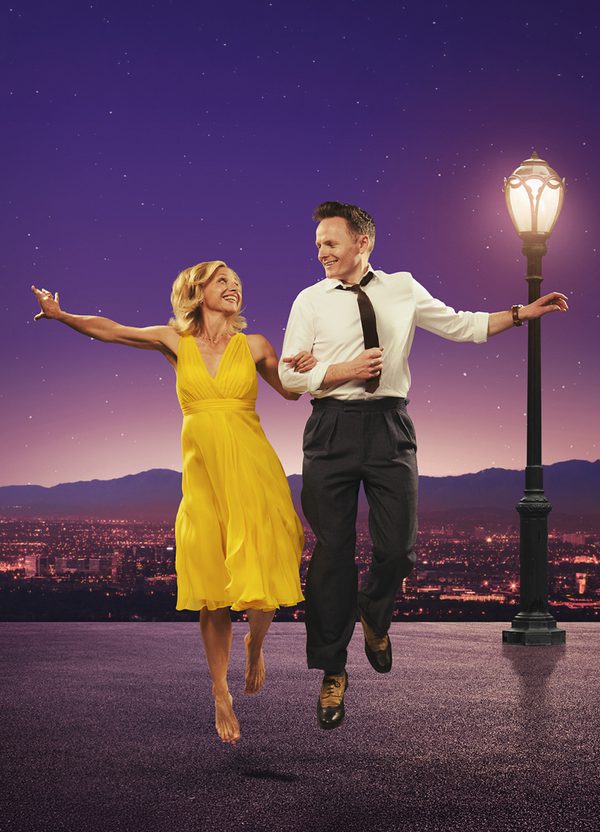 In a tribute to the film poster for La La Land, Liza Pulman and Joe Stilgoe dance together against a purple-hued vista of Hollywood, with a lighted street lamp beside them. She wears a sleeveless yellow dress, he is in white shirt, black tie and trousers, and they smile happily at each other as they leap off the ground.