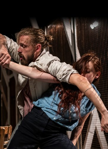 A man and woman entwined in a dance pose. The bearded man's hair is tied back and he wears a white jacket with rolled up sleeves. The woman has long brown hair; her face is partially obscured by the man's arm and she wears a sleeveless blue shirt. Behind them is a wire mesh structure.