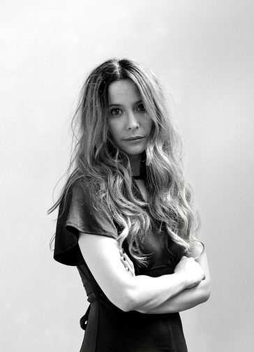 A black and white photo of a woman with long blonde hair cascading down past her shoulders, wearing a short-sleeved black dress. Her arms are folded and she looks at the camera with a slight smile.