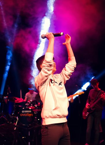 A young boy wearing a white hoodie raises his arms aloft, with a microphone in his left hand. A drummer and guitarist are playing behind him, illuminated by smoky pink lights and white lasers.