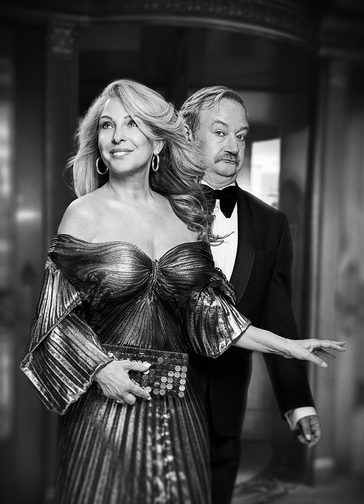 A black and white photo of a glamorous blonde woman in an off-the-shoulder pleated lurex dress and big gold hoop earrings, with a confident smile. Behind her a man with a moustache, wearing formal black tie, looks towards the camera with an exasperated expression.