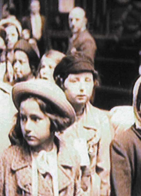 A grainy historical black and white photo of a group of children in the 1930s, dressed in coats and hats, some wearing labels around their necks and looking anxious and uncertain.