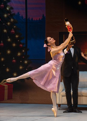 A female dancer wearing a pink satin party dress, her dark hair fastened in a bun with a pink bow, performs an arabesque. She's holding a Nutcracker doll aloft. Behind her is a decorated Christmas tree.