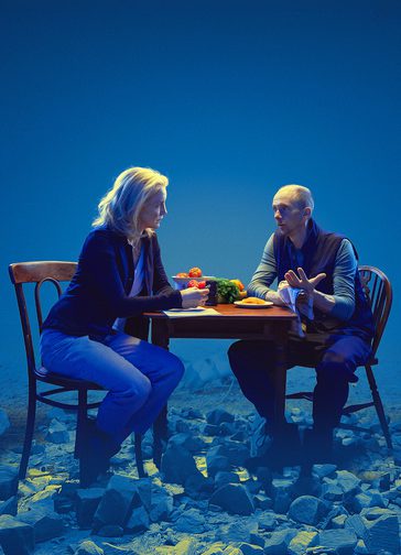 A blonde woman and a short-haired man sit looking at each other, in serious conversation, on either side of a table on which are bowls and plates of tomatoes and other fruit and vegetables. She has a notebook. They are illumined against a blue background and the ground is covered in shattered rocks.