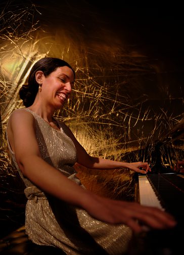 A smiling woman, her dark hair tied back in a chignon, sits playing the piano. She wears a sleeveless, dull gold shimmering dress, and has an illuminated gold background behind her.
