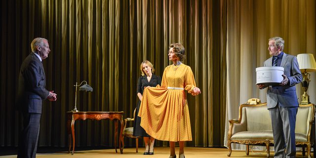 A woman in a 1960s yellow dress and black heels stands in the middle of the stage. She holds the side of the dress out to the side of her and looks at a man in a checked suit expectantly. A woman in a black skirt suit stands behind, also looking at the man expectantly. A man to the right of the woman in yellow holds a large hat box, dressed in a grey suit. He raises his eyebrows at the other man. Behind them is a delicate writing desk and lamp and a gold detailed sofa.