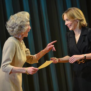 A woman in a beige three-quarter length sleeve dress and short curled grey hair holds a piece of paper in one hand and points slightly at the woman next to her. She is smiling. The other woman is dressed in a black skirt suit and smiles back at the woman.