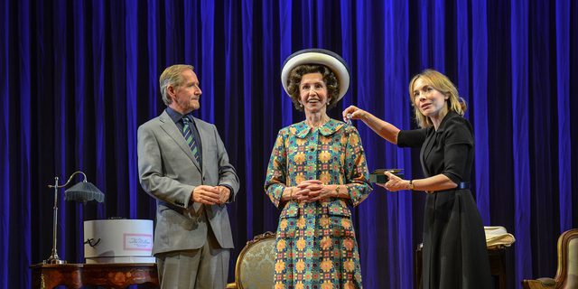 Three actors stands on stage, two women looking forward as if they were looking into a mirror. The woman in the middle wears a bright ecclectic patterned dress and a wide brimmed hat. She smiles widely, her hands knitted in front of her. The woman on the right in a black skirt suit holds up a brooch in front of the woman in the middle, seeing what it would look like on. The man on the left looks on at the two women.