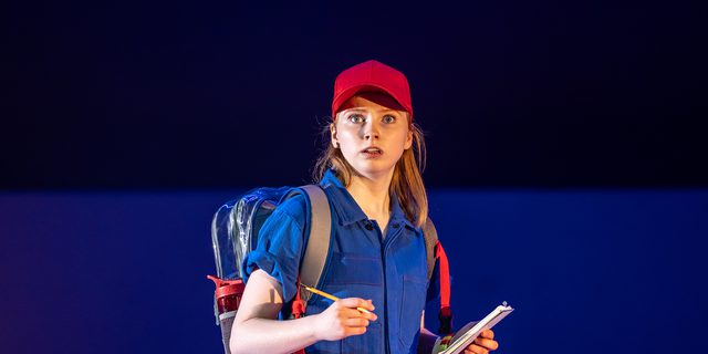 A woman in a blue short-sleeve jumpsuit and a red cap and clear rucksack holds a notepad in one hand and a pencil in the other. She looks off, worried or shocked, her mouth slightly open.