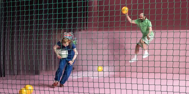 Behind a green net, a man is jumping mid-air. He holds a yellow ball above his head, moving as if he were to throw it at the woman in front of him. Other yellow balls litter the pink stage. The woman is wearing a blue short-sleeve jumpsuit and is wearing a large rucksack and holds an animal cage under one arm. She is ducking to avoid being hit.