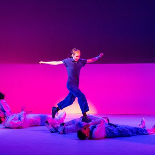 A woman in a short-sleeved jumpsuit and boots is mid-jump. She is jumping over other members of the company as they roll underneath her. They are all wearing matching tracksuits. Her arms are outstretched and has a look of concentration on her face. A bright pink washes covers the stage.