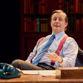 A man in a striped shirt, blue striped tie, red braces and pin-striped trousers sits behind a desk. He is leaning back in his chair, with his suit jacket over the back of it. On the desk is a teal telephone, a wooden box and piles of paper. He has one hand hovering just above the table.