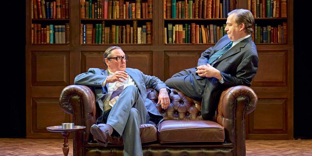 Two men sit on a leather sofa. The man on the left is wearing a grey checked suit, glasses and slicked over hair. He has one leg crossed over the other and his arms draped over the back and arm of the sofa. He is holding a glass in his hand. He is raising his eyebrows at the man on the right who is sat on the back of the sofa, leaning backwards onto the arm of the sofa. He is wearing a dark pin-striped suit. His hands are clasped together. Behind is a line of bookshelves and has wooden parquet flooring.
