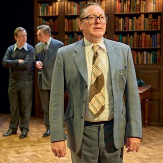 A man in a grey checked suit, a brown and white tie, glasses and slicked over hair stands with his hands by his side. He looks into the distance, his mouth slightly apart. Two men stand behind him, one man in a shirt and jumper with his arms crossed, the other in a suit and holding a wine glass. They are looking at each other, in mid conversation. They are stood in a library, with a back wall of bookshelves and wooden parquet flooring.