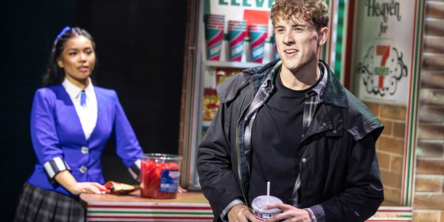 A teenage boy dressed in black wax jacket, dark checked shirt and black tee shirt, stands holding a colourful sports disposable cup. He has short curly brown hair and is smiling. On the left, slightly out of focus, a girl with brown hair tied back in a ponytail, wearing a close fitting purple blazer and short tartan skirt, stands next to a shop counter on which is displayed confectionary; behind it are 7-Eleven shop signs.