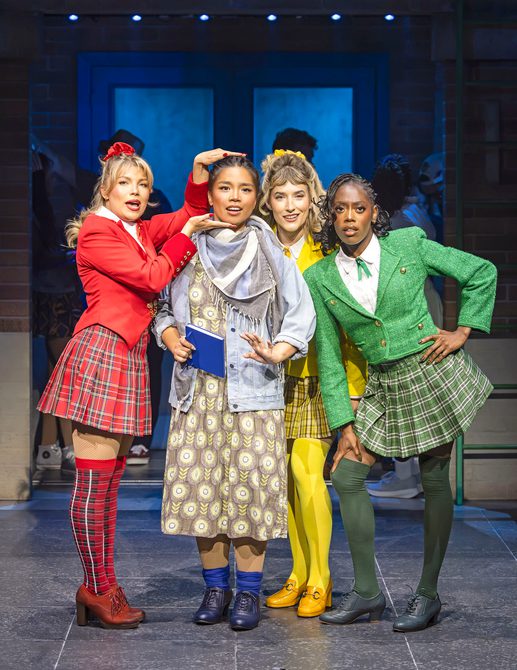 Four girls stand facing the camera. In the centre a girl in a long printed dress, shapeless jacket, scarf and rolled down ankle socks, holds a blue notebook. She's surrounded by three girls in brightly coloured close-fitting blazers, tartan mini skirts and matching knee high socks who stare as though comparing their reflections in a mirror.