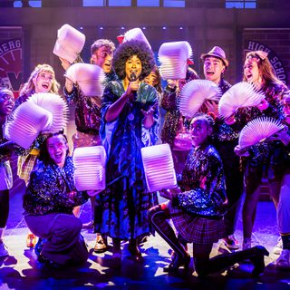 A large group of performers surround a woman with dark curly hair who stands singing in the centre, holding a microphone. All the ensemble are smiling broadly and wear sparkly jackets and hold open books whose pages resemble pompoms.