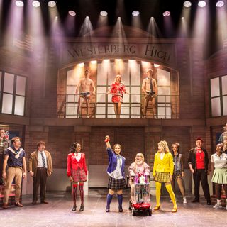 A group of performers are pictured in the school hall of Westerberg High. On an upper balcony in front of the window is a blonde girl in a red mini dress, with two young men in briefs and a tie. Below them, girls, women and men stand singing in a semi-circle. In the centre, a girl using a mobility scooter has three girls in blazers, tartan mini skirts and knee-high socks - respectively red, purple and yellow - either side of her.