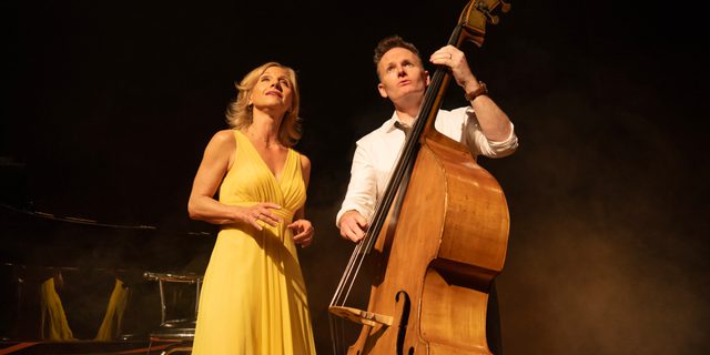 A man and a woman stand side by side, looking upwards. The woman has blonde hair and wearing a long sleeveless yellow dress. The man is in a white shirt with his sleeves rolled up, holding and playing a double bass.