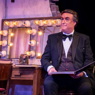 A man in black tie sits in a dressing room with an open book on his lap.