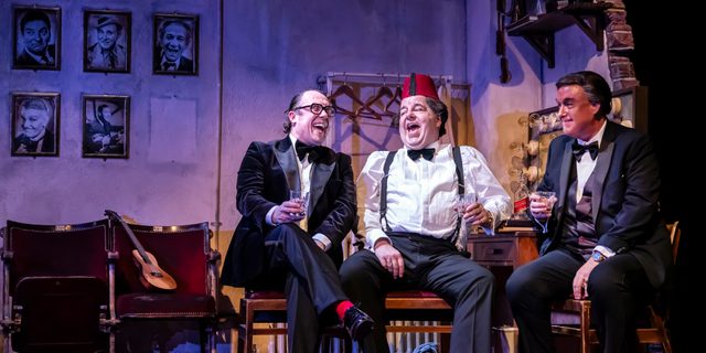 Three comedians laugh in a dressing room. They are all in formal clothing, the man in the middle wears a fez.