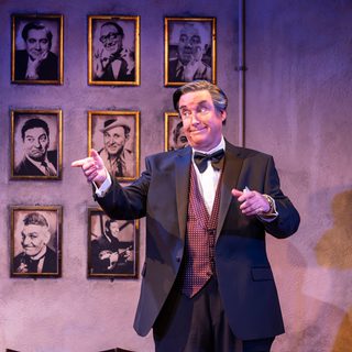 A man in a black suit and polka dot waist coat stands in front of a wall with portraits of men in black and white. He points with one hand, his eyebrows raised and mouth in a closed smile.