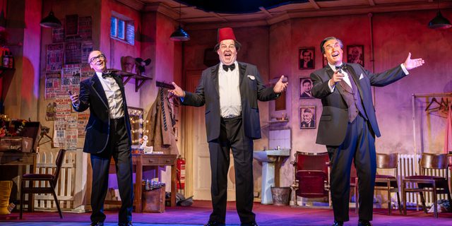 Three men stand in a theatre dressing room, the arms stretched out in front of them. They all wear formal suits, the man in the middle wears a red fez. They all have their mouths open and smiling, as if singing. The back of the stage is washed in a pink-red hue.