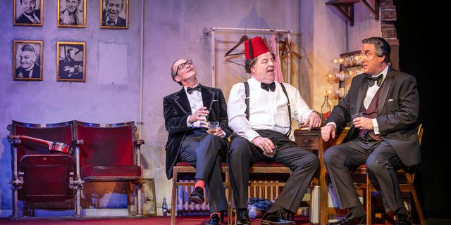 Three men sit in a theatre dressing room. Gold frames with male comedians hang on the wall. The men are dressed in suits. The man in the middle wears a red fez.